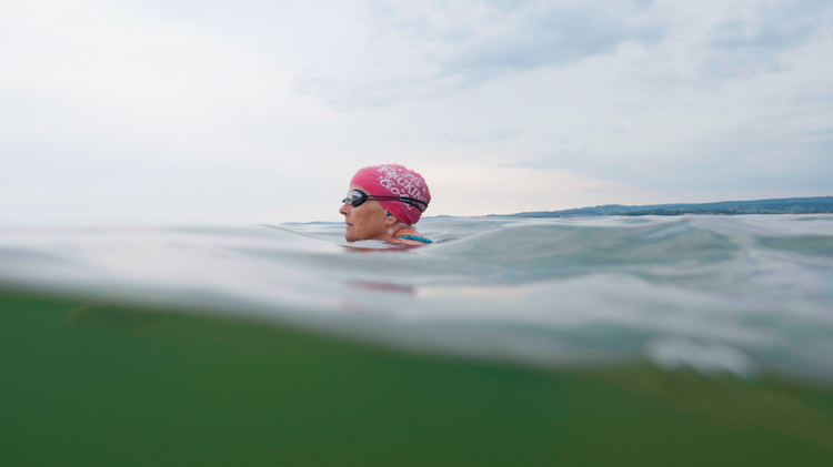 Swimmer wearing a pink swim cap in open water under a cloudy sky with distant shoreline in the background.