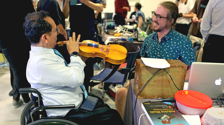 A person playing a bass attached to electronic contraptions with an audience watching.