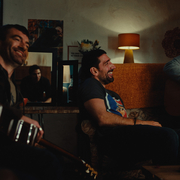 A group of people playing acoustic guitars and sitting in a cozy, dimly lit living room with warm décor.