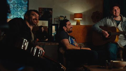 A group of people playing acoustic guitars and sitting in a cozy, dimly lit living room with warm décor.