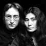 Black and white portrait of two people with long hair, wearing dark shirts, close together against a black background.