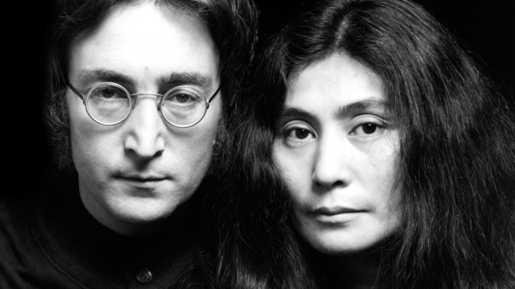 Black and white portrait of two people with long hair, wearing dark shirts, close together against a black background.