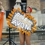 Two people in swimwear at an outdoor event, one holding a sign reading "Rave on for the Avon" near a pool and flowerbed.
