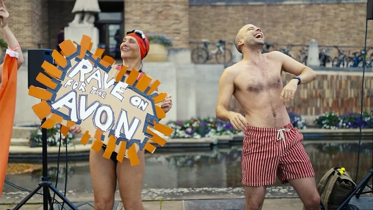 Two people in swimwear at an outdoor event, one holding a sign reading "Rave on for the Avon" near a pool and flowerbed.
