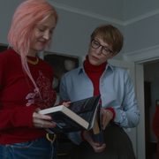 Three people indoors, two are looking at books while the third stands in the background near a doorway.