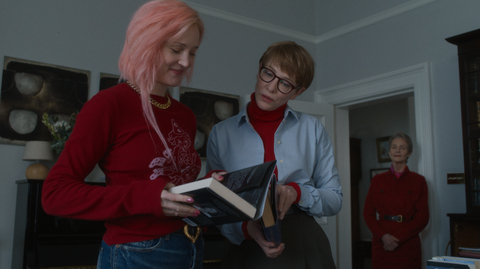 Three people indoors, two are looking at books while the third stands in the background near a doorway.