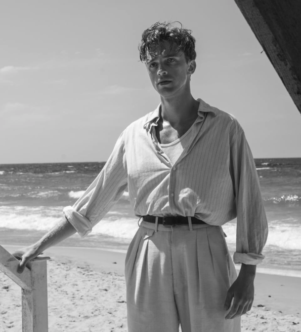 Black and white photo of a person standing on a beach, leaning on a wooden railing with waves in the background.