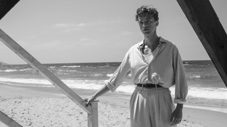 Black and white photo of a person standing on a beach, leaning on a wooden railing with waves in the background.