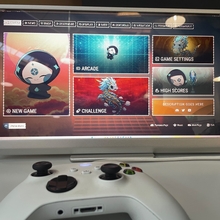 A gaming monitor displays a colourful game menu with options like New Game, Arcade, and Challenge. A white controller lies in the foreground, ready for use.