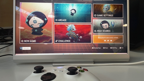 A gaming monitor displays a colourful game menu with options like New Game, Arcade, and Challenge. A white controller lies in the foreground, ready for use.