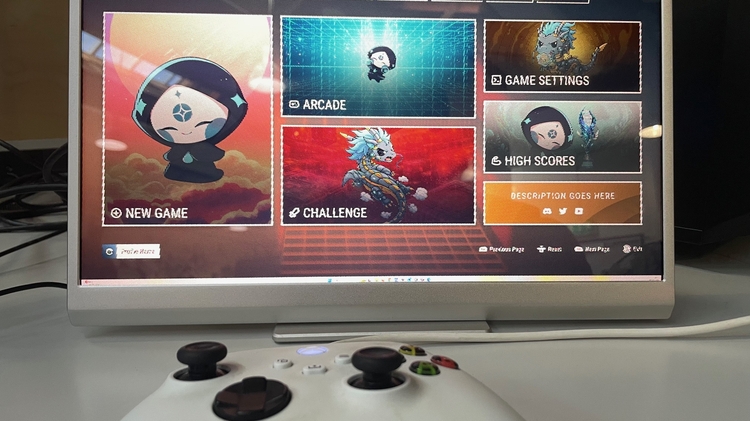 A gaming monitor displays a colourful game menu with options like New Game, Arcade, and Challenge. A white controller lies in the foreground, ready for use.