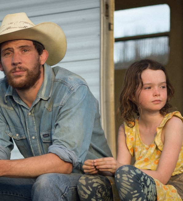 A man in a cowboy hat and denim shirt sits beside a young girl in a yellow dress outside a trailer home.
