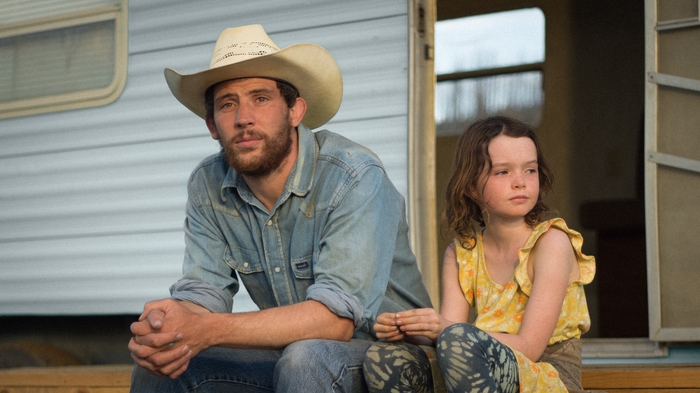 A man in a cowboy hat and denim shirt sits beside a young girl in a yellow dress outside a trailer home.