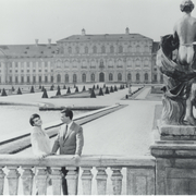 Black and white photo of a couple by a balustrade, with a grand palace and manicured gardens in the background, and a stone statue nearby.