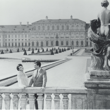 Black and white photo of a couple by a balustrade, with a grand palace and manicured gardens in the background, and a stone statue nearby.