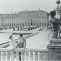 Black and white photo of a couple by a balustrade, with a grand palace and manicured gardens in the background, and a stone statue nearby.