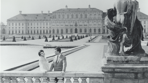 Black and white photo of a couple by a balustrade, with a grand palace and manicured gardens in the background, and a stone statue nearby.