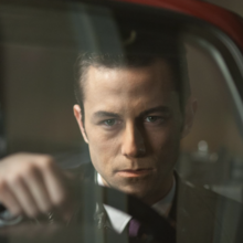 Person holding steering wheel inside a car at night, viewed through the windshield with a passenger's arm visible.