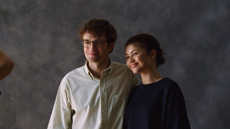 A man in a white shirt and a woman in a black jumper stand next to each other and smile looking away from the camera.