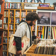 An image of the musician Lawi Anywar a Black man with an afro is browsing vinyl records in a vintage market. He is surrounded by various types of physical media, and carries a white tote bag full of records. 