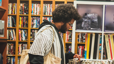 An image of the musician Lawi Anywar a Black man with an afro is browsing vinyl records in a vintage market. He is surrounded by various types of physical media, and carries a white tote bag full of records. 