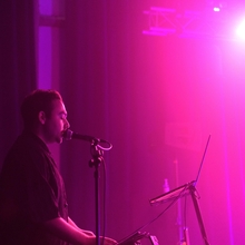 A musician performs on stage under vibrant purple lighting, singing into a microphone. He stands by a keyboard with a focused expression, creating an immersive atmosphere.