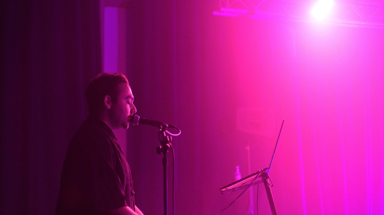 A musician performs on stage under vibrant purple lighting, singing into a microphone. He stands by a keyboard with a focused expression, creating an immersive atmosphere.