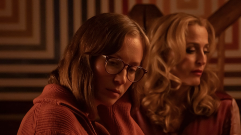 Two people in warm lighting with one wearing an orange sweater and the other with curly blonde hair, against a geometric patterned background.