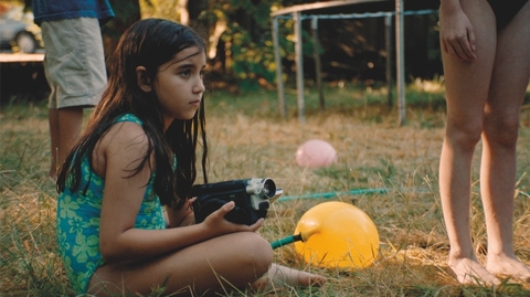 Child in a blue swimsuit sitting on grass holding a vintage video camera, nearby yellow and pink balloons and barefoot people standing.
