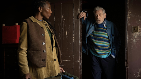 Two people standing inside a dimly lit room with worn brown wooden doors, wearing layered cozy clothing.