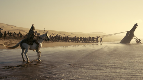 A soldier on a white horse watches as a large group pulls a giant statue from the water onto a sandy shore at sunset.