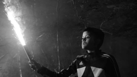 A boy in a dark jacket holding a lit flare in a dim forest setting at night.