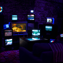 An image of a darkened room, illuminated only by a series of CRT TV's organised in a disparate fashion, all activated and showing different images, creating a collage of sorts. There is a sofa placed in front of the installation, for the viewer to experience it. 