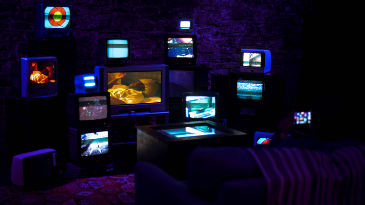 An image of a darkened room, illuminated only by a series of CRT TV's organised in a disparate fashion, all activated and showing different images, creating a collage of sorts. There is a sofa placed in front of the installation, for the viewer to experience it. 
