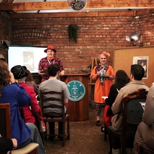 An image depicting Paul (on the left in red) and Gill (on the right in orange) delivering a theatre experience in a red brick room with a smartly dressed audience. They are flanked by a cork board on the right and a small projection set upon the left. 