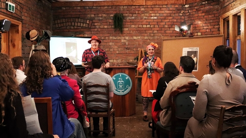 An image depicting Paul (on the left in red) and Gill (on the right in orange) delivering a theatre experience in a red brick room with a smartly dressed audience. They are flanked by a cork board on the right and a small projection set upon the left. 