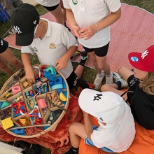 An image of young people gathered around a container of craft materials as they deliberate how to use them. Two of them are sat down on an orange covered seat on the right, and four more stand in the center of the image, interfacing with the container of craft materials. 