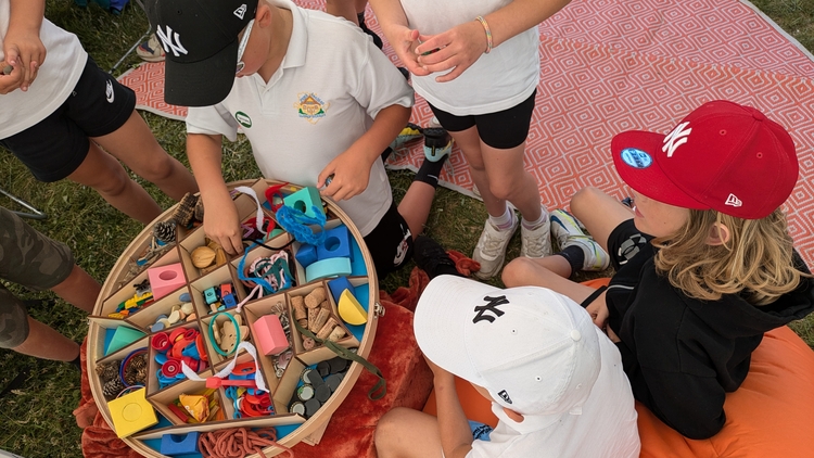 An image of young people gathered around a container of craft materials as they deliberate how to use them. Two of them are sat down on an orange covered seat on the right, and four more stand in the center of the image, interfacing with the container of craft materials. 