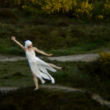 An image of Anastasia Dahl, dressed entirely in white, performing a piece of movement in an outdoor earthy green space - she is surrounded by yellow flowering gorse bushes to the top and right of the image. She is stood on one leg, with her left arm and leg pointing outward, and her right arm pointing upward. Her head faces the right of the image. 