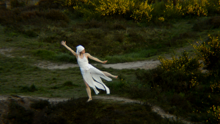 An image of Anastasia Dahl, dressed entirely in white, performing a piece of movement in an outdoor earthy green space - she is surrounded by yellow flowering gorse bushes to the top and right of the image. She is stood on one leg, with her left arm and leg pointing outward, and her right arm pointing upward. Her head faces the right of the image. 