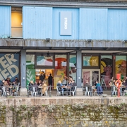 Undershed with people dining under a blue building featuring large Watershed sign and colourful mural panels.