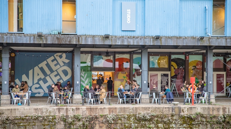 Undershed with people dining under a blue building featuring large Watershed sign and colourful mural panels.