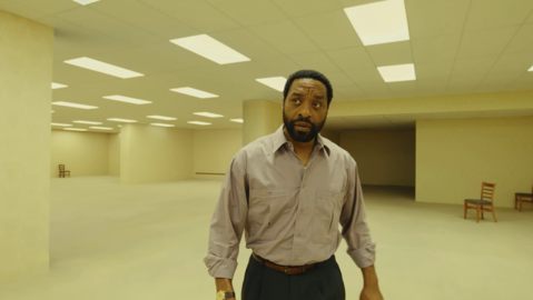 A man in a beige shirt and dark trousers stands in a large, empty, yellow-toned room with scattered wooden chairs and ceiling lights.