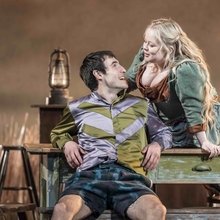 Two actors in period costumes engage on a rustic set with wooden furniture and a lantern.
