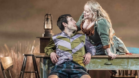 Two actors in period costumes engage on a rustic set with wooden furniture and a lantern.