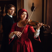 Woman in red Renaissance dress playing violin indoors, with a man in dark period attire standing behind her near hanging violins.