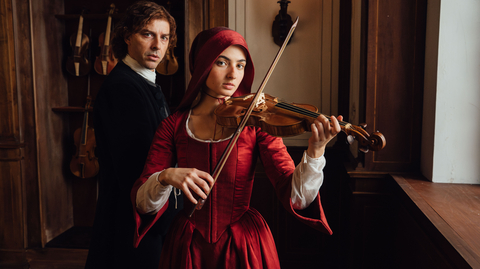 Woman in red Renaissance dress playing violin indoors, with a man in dark period attire standing behind her near hanging violins.