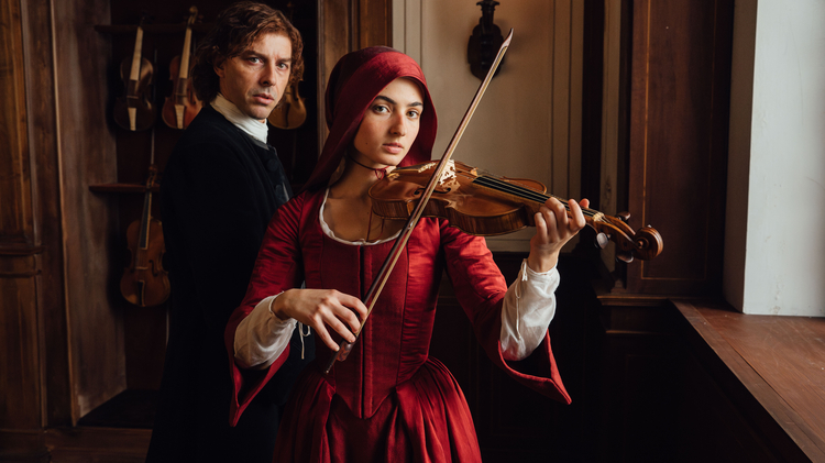 Woman in red Renaissance dress playing violin indoors, with a man in dark period attire standing behind her near hanging violins.