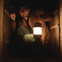 Person crouching in a small dark wooden space, holding a lit lantern glowing warmly in the dim surroundings.