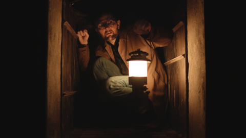 Person crouching in a small dark wooden space, holding a lit lantern glowing warmly in the dim surroundings.